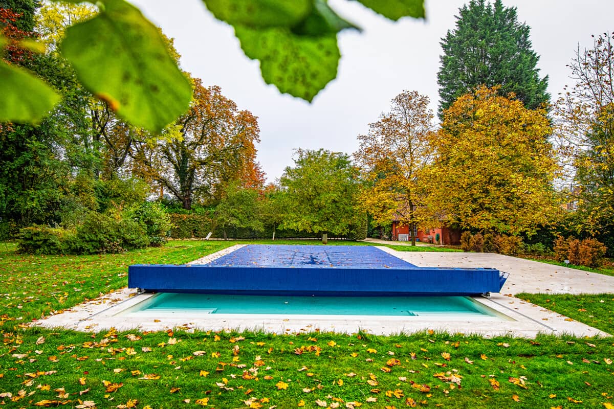 Piscine couverte d'une bâche bleue dans un jardin entouré d'arbres aux feuilles automnales, avec une maison en arrière-plan.