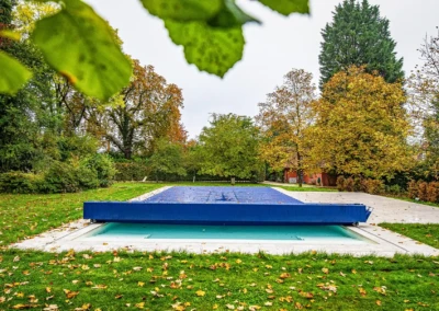 Piscine couverte d'une bâche bleue entourée de pelouse et d'arbres aux feuilles automnales.