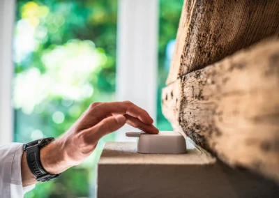 Een hand met een zwart horloge drukt op een witte behuizing die op een oppervlak ligt in de buurt van een houten balk, tegen een groene achtergrond.