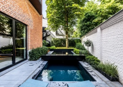 Chez-Anna-et-Luc-1 Kleine rechthoekige zwembad omringd door muren van rode en witte bakstenen, met groene struiken en een grote boom op de achtergrond.