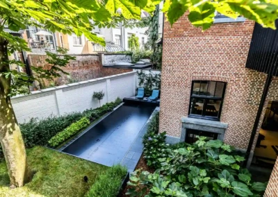 Chez-Anna-et-Luc-3 Een tuin met een smal zwembad omgeven door groen, twee blauwe ligstoelen en een muur van rode bakstenen.