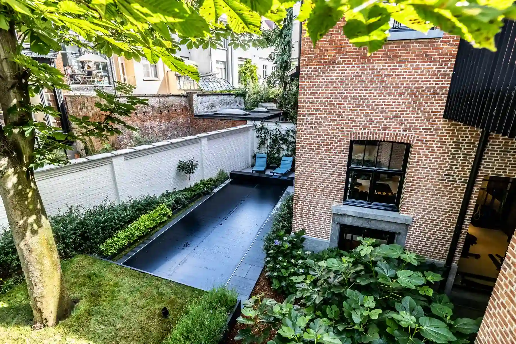 Cour arrière avec piscine couverte noire, entourée de murs en briques rouges et de végétation, deux chaises longues bleues visibles.