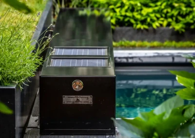 Chez-Anna-et-Luc-4 Zwarte behuizing met zonnepanelen naast een zwembad, omringd door groene planten.