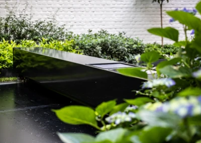 Chez-Anna-et-Luc-6 Een rechthoekige zwarte tafel buiten, omringd door groene planten en blauwe bloemen, met een witte bakstenen muur op de achtergrond.
