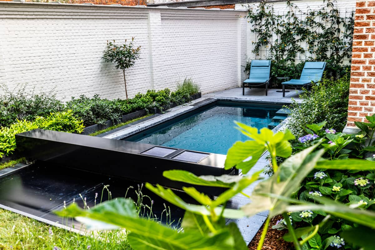 Kleine zwembad omringd door groen met twee blauwe ligstoelen op een betonnen terras, omzoomd door muren van witte bakstenen.