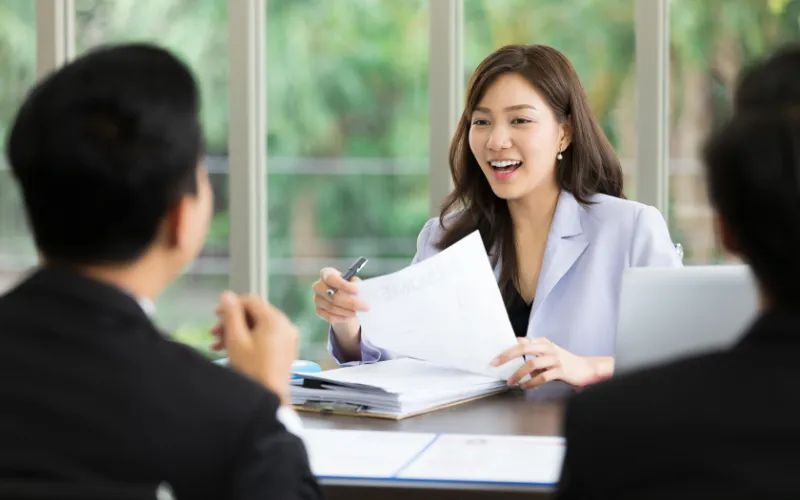 Een glimlachende vrouw in een grijs pak houdt documenten vast tegenover twee zittende personen, in een helder kantoor met ramen.