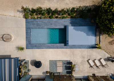 Luchtfoto van een rechthoekig zwembad omgeven door grijze tegels, met ligstoelen en een ronde zwarte tafel op een betonnen terras.