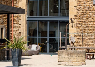 Stenen gevel met groot rond raam, terras met grijze fauteuils, stenen put en plant in zwarte pot.