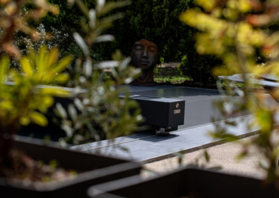 Een gezicht, gebeeldhouwd in brons, is omgeven door vegetatie, met een metalen structuur op de voorgrond op een grijze terras.