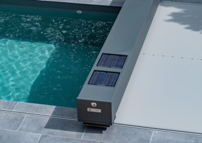 Grijze zwembadhoes met geïntegreerde zonnepanelen, omringd door grijze tegels en vegetatie op de achtergrond.