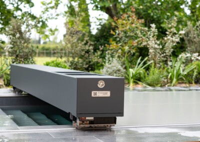 Een rechthoekig, metalen grijs apparaat is geplaatst aan de rand van een zwembad, omgeven door weelderig groen.