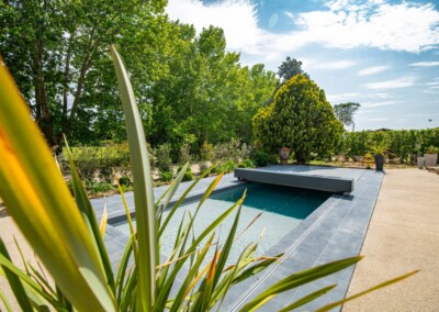 Een zwembad met een grijze hoes is omringd door weelderige vegetatie en potplanten onder een gedeeltelijk bewolkte hemel.