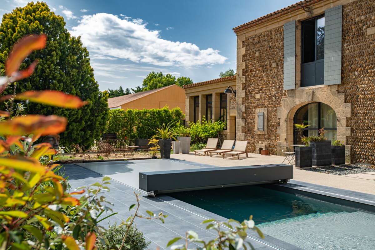 Een zwembad met een grijze afdekking wordt omringd door groen, vlakbij een stenen huis met ligstoelen en potplanten.