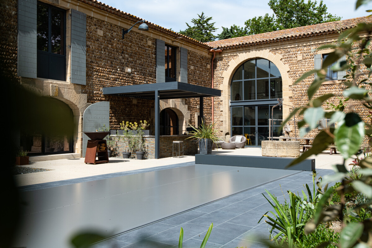 Cour intérieure avec façade en pierre, pergola grise, mobilier de jardin et couverture de piscine grise entourée de plantes vertes.