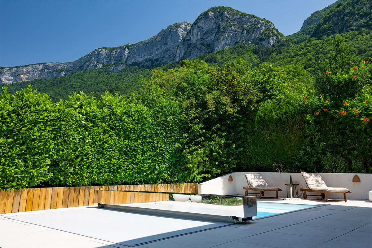 Binnenzwembad bedekt met een rolluik, omringd door ligstoelen en weelderige vegetatie, met bergen op de achtergrond onder een blauwe hemel.