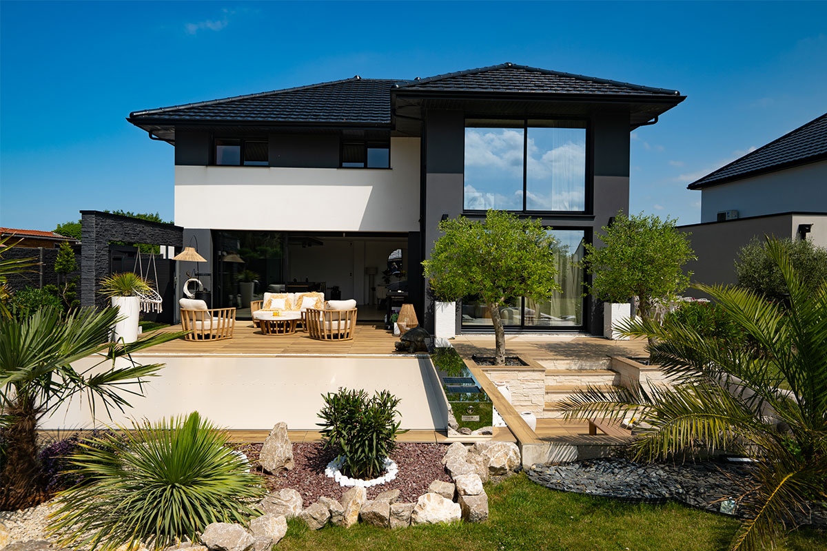 Moderne woning met houten terras, beige tuinmeubilair, groene planten en bomen, onder een helderblauwe lucht.