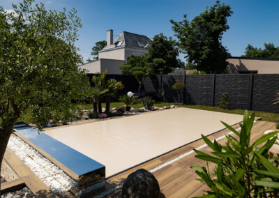 Een tuin met een beige overdekt zwembad, omringd door groene planten en witte stenen, voor een modern huis.