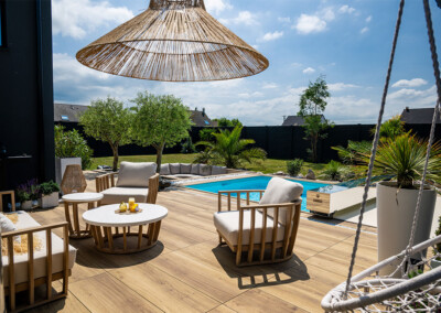 Houten terras met witte fauteuils, lage tafel en zwembad omgeven door groen onder een blauwe hemel.