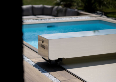 Een glanzende metalen zwembadafdekking is geïnstalleerd aan de rand van een zwembad omringd door witte stenen en licht hout.