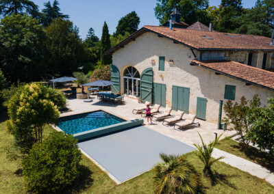 Groot stenen huis met zwembad, omringd door weelderige vegetatie en ligstoelen op een zonnig terras.