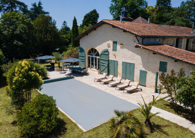 Groot stenen huis met groene luiken, zonnig terras, ligstoelen en overdekt zwembad, omgeven door weelderige vegetatie.