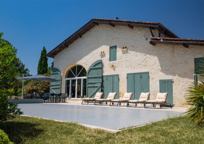 Stenen huis met groene luiken, omgeven door een tuin, heeft een terras met ligstoelen en een eettafel onder een parasol.