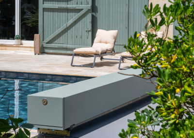 Twee beige ligstoelen bij een zwembad, met een grijze rolluik en een groene muur op de achtergrond.