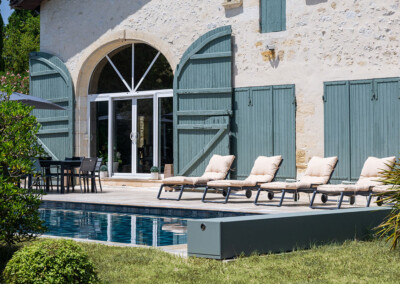 Beige ligstoelen voor een stenen huis, met groene luiken en een zwembad op de voorgrond.