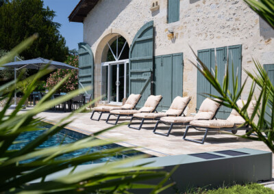 Beige ligstoelen op een stenen terras bij een zwembad, voor een huis met groene luiken en een grijze parasol.
