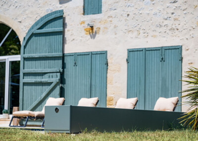 Gevel van een stenen huis met groene luiken, beige ligstoelen en een gazon op de voorgrond, onder een heldere hemel.