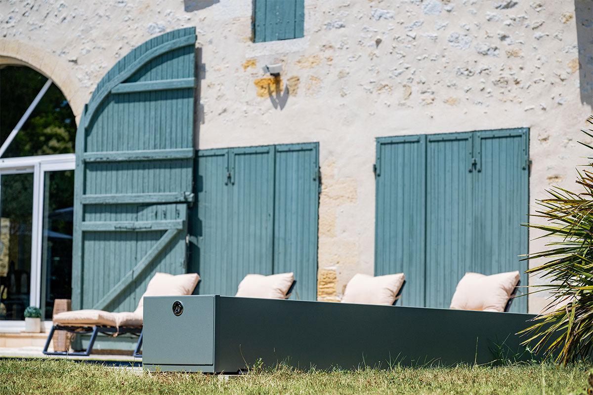 Gevel van een stenen huis met groene luiken, beige ligstoelen en een gazon op de voorgrond, onder een heldere hemel.
