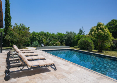 Beige ligstoelen uitgelijnd op een stenen terras grenzen aan een rechthoekig zwembad omgeven door weelderige vegetatie onder een blauwe hemel.